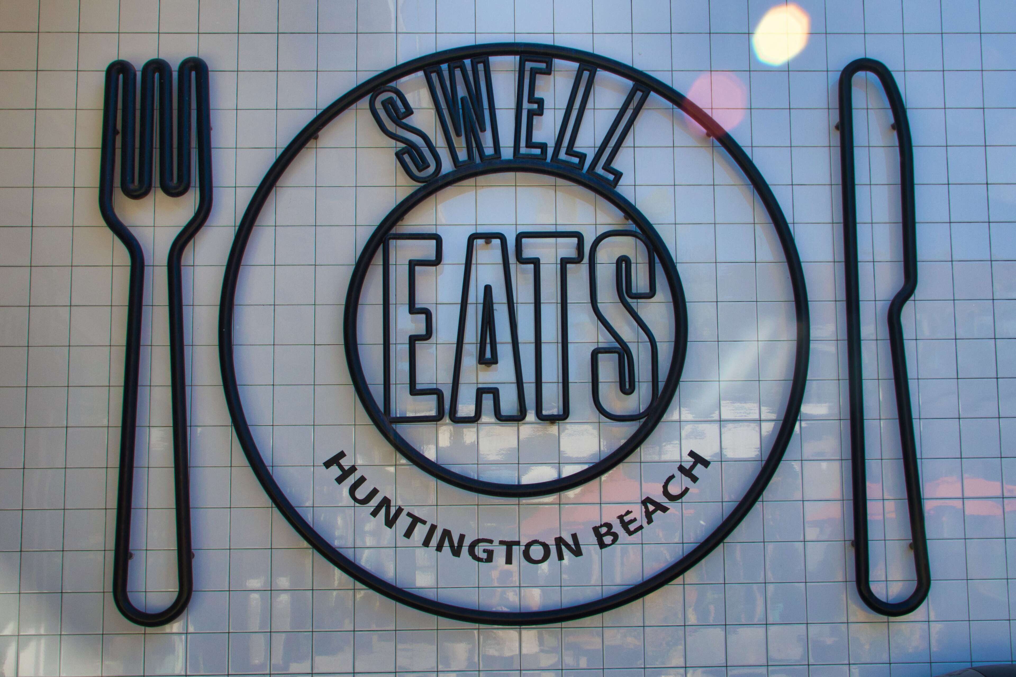A sign with the words "SWELL EATS" and "HUNTINGTON BEACH" on a tiled wall.