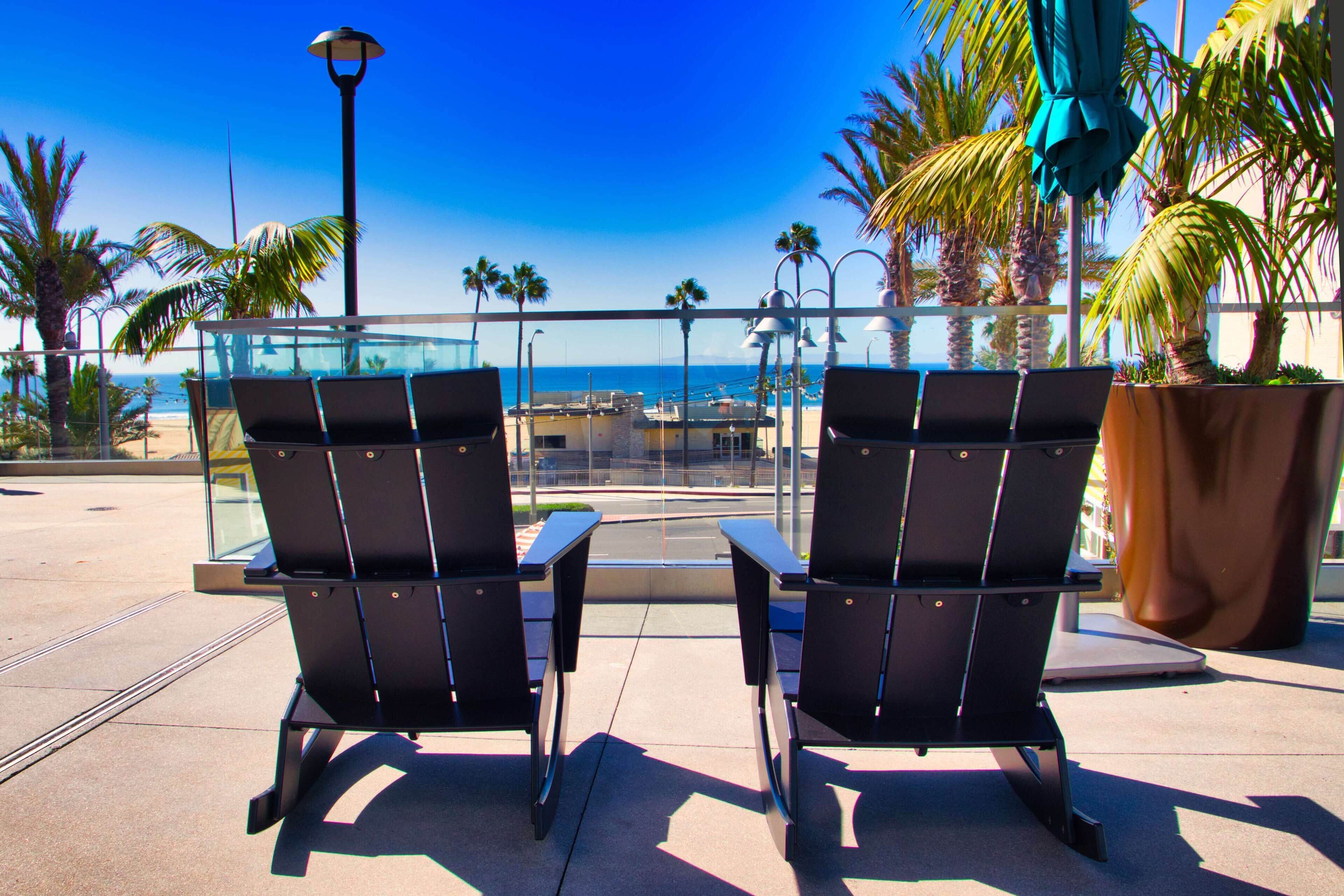 Two black chairs with metal frames and a glass table, overlooking a beach with palm trees and a clear blue sky.
