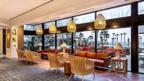 A spacious lobby with wicker chairs, a leather sofa, and large windows offering a view of palm trees and a beach. - The Waterfront Beach Resort, A Hilton Hotel (Huntington Beach)