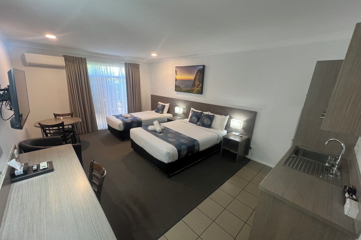 A hotel room with two beds, a desk, a chair, a small table, a TV, a painting on the wall, and a sink.