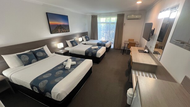 Une chambre d’hôtel avec trois lits, un bureau, une chaise, une télévision et une fenêtre avec des rideaux.