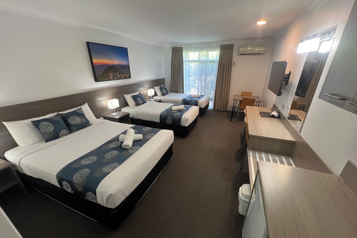 A hotel room with three beds, a desk, a chair, a TV, and a window with curtains.