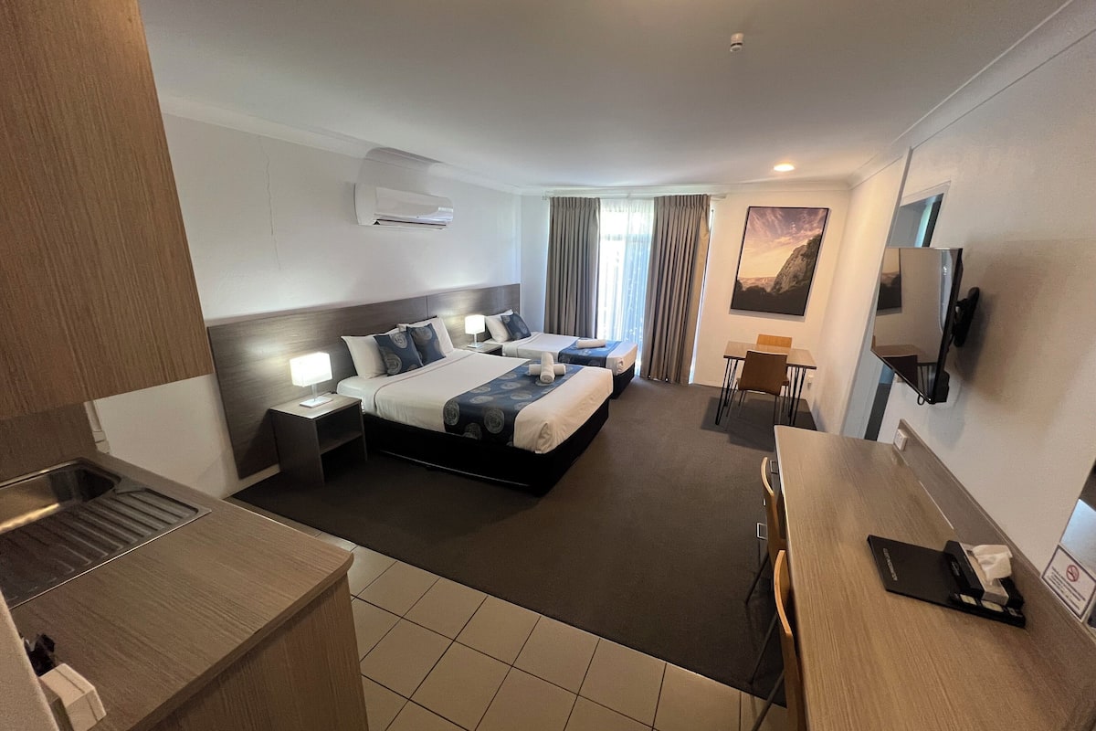 A hotel room with a large bed, a desk, a chair, a TV, and an air conditioning unit.