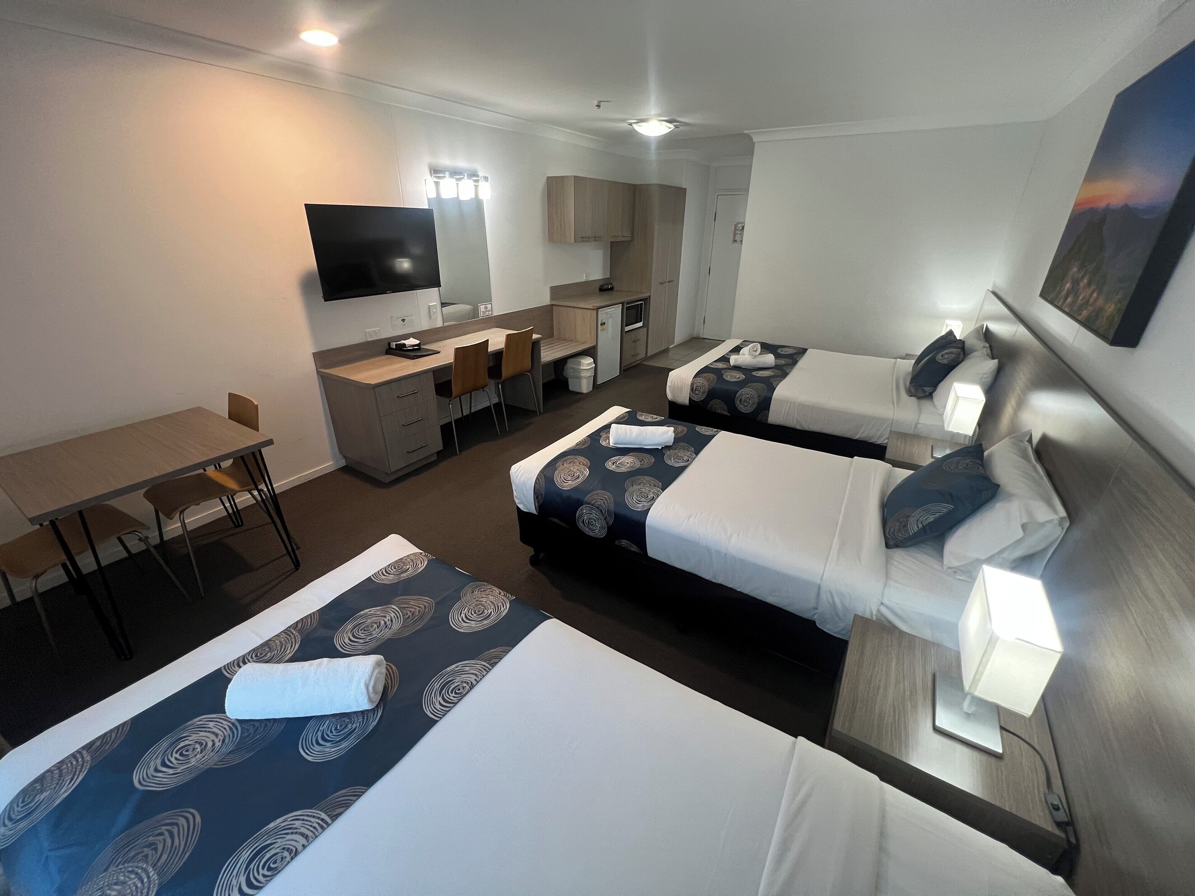 A hotel room with two beds, a desk, a chair, a TV, and a small kitchenette.