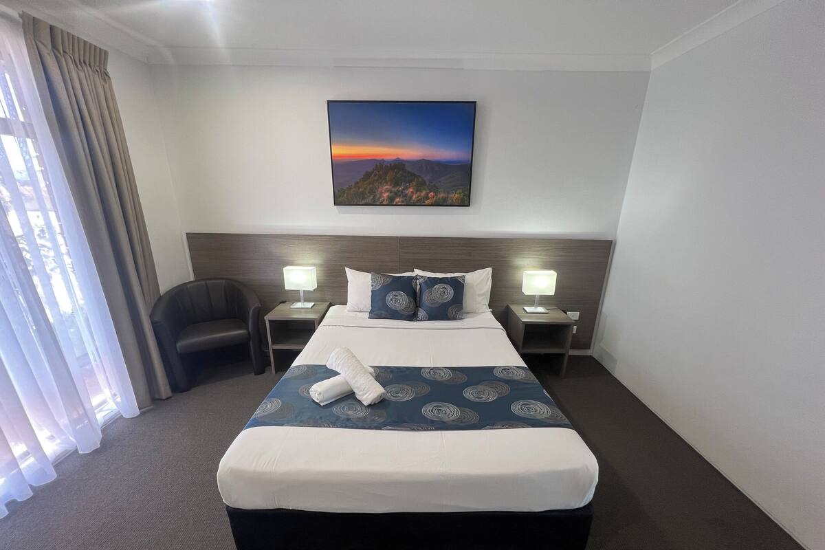 A hotel room with a large bed, two bedside tables with lamps, a chair, a painting on the wall, and a view of the outdoors through the window.