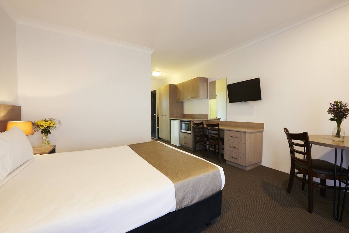 A hotel room with a bed, desk, kitchenette, and a TV mounted on the wall.