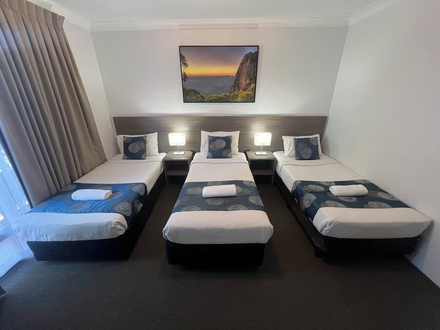 Ett hotellrum med tre enkelsängar, var och en med en kudde och ett täcke. Mellan sängarna står ett nattduksbord med en lampa. En tavla med ett landskapsmotiv hänger på väggen ovanför sängarna.