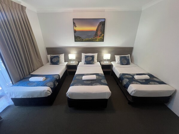 A hotel room with three single beds, each with a pillow and a blanket. There is a nightstand with a lamp between the beds. A painting of a landscape is on the wall above the beds.