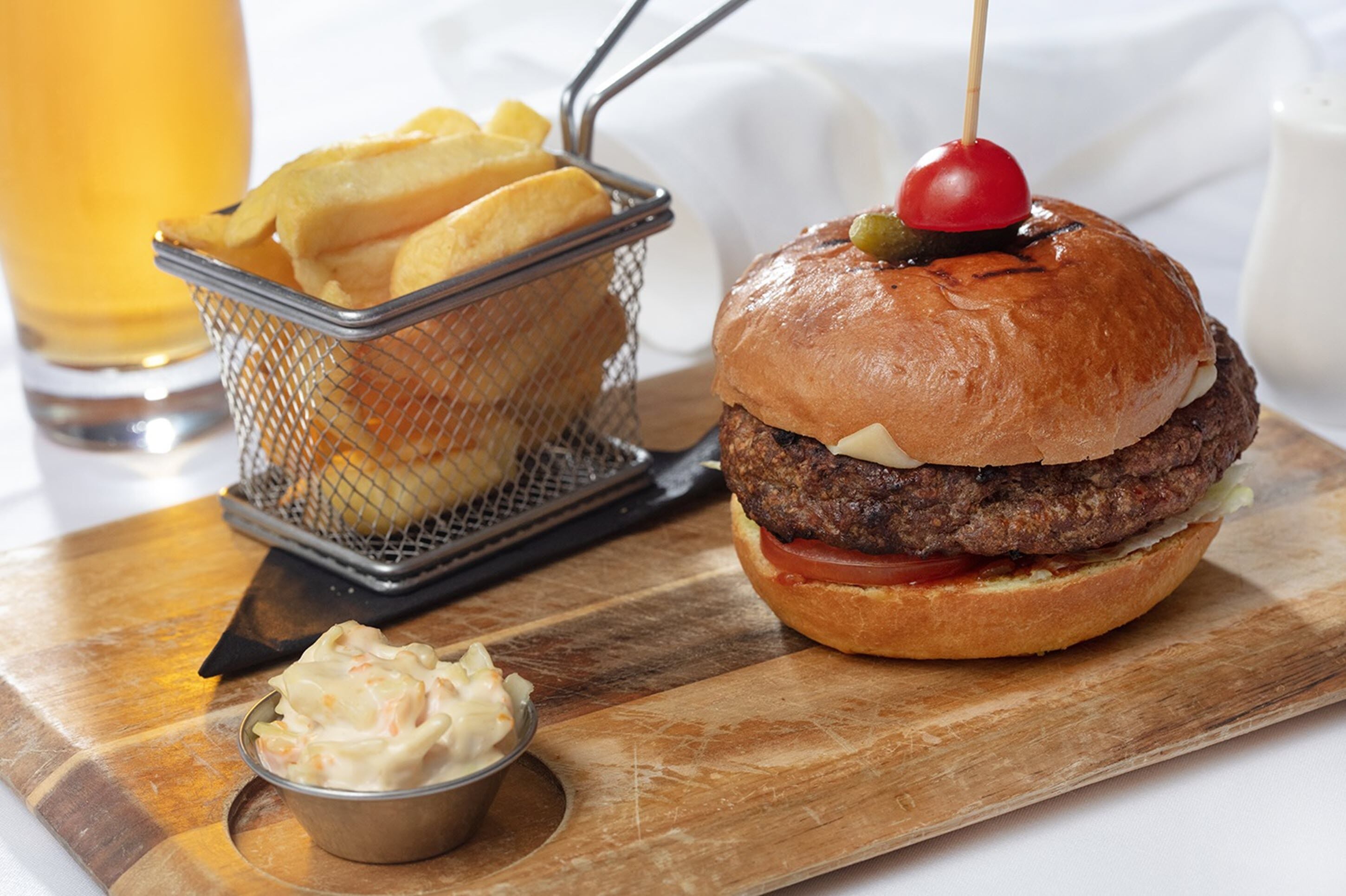 A burger with a cherry on top, a basket of fries, and a small bowl of sauce on a wooden board.