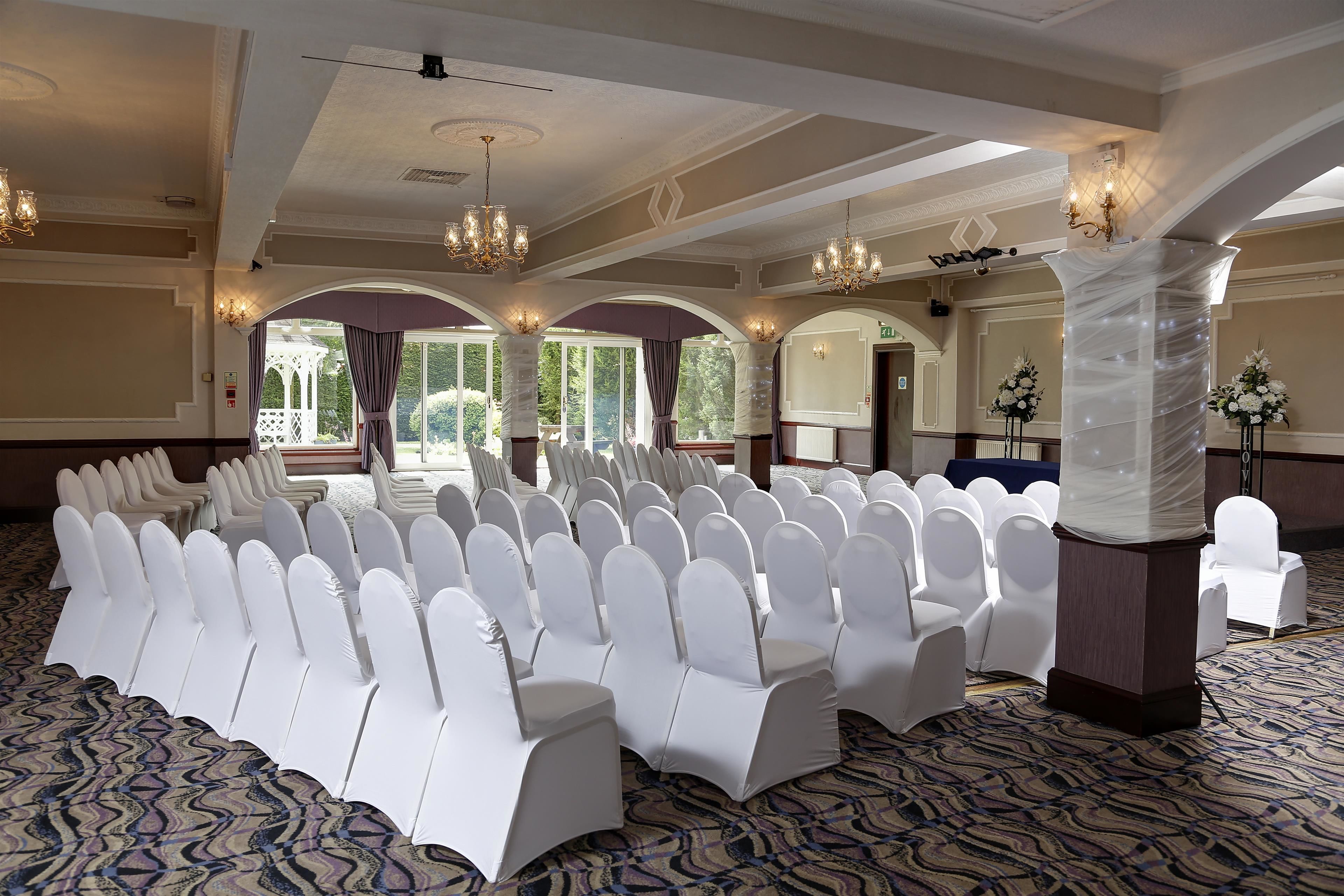 A banquet hall with rows of white chairs and tables, chandeliers, and a patterned carpet.