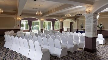 A banquet hall with rows of white chairs and tables, chandeliers, and a patterned carpet.