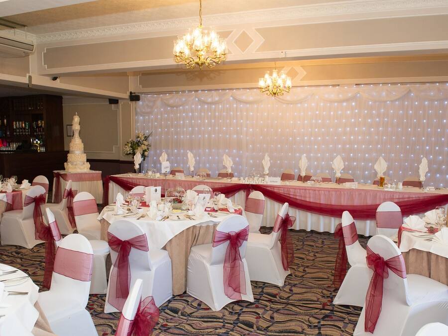 Un salón de banquetes con mesas redondas dispuestas para un evento formal, con manteles blancos y rosados, y sillas adornadas con cintas rosas.