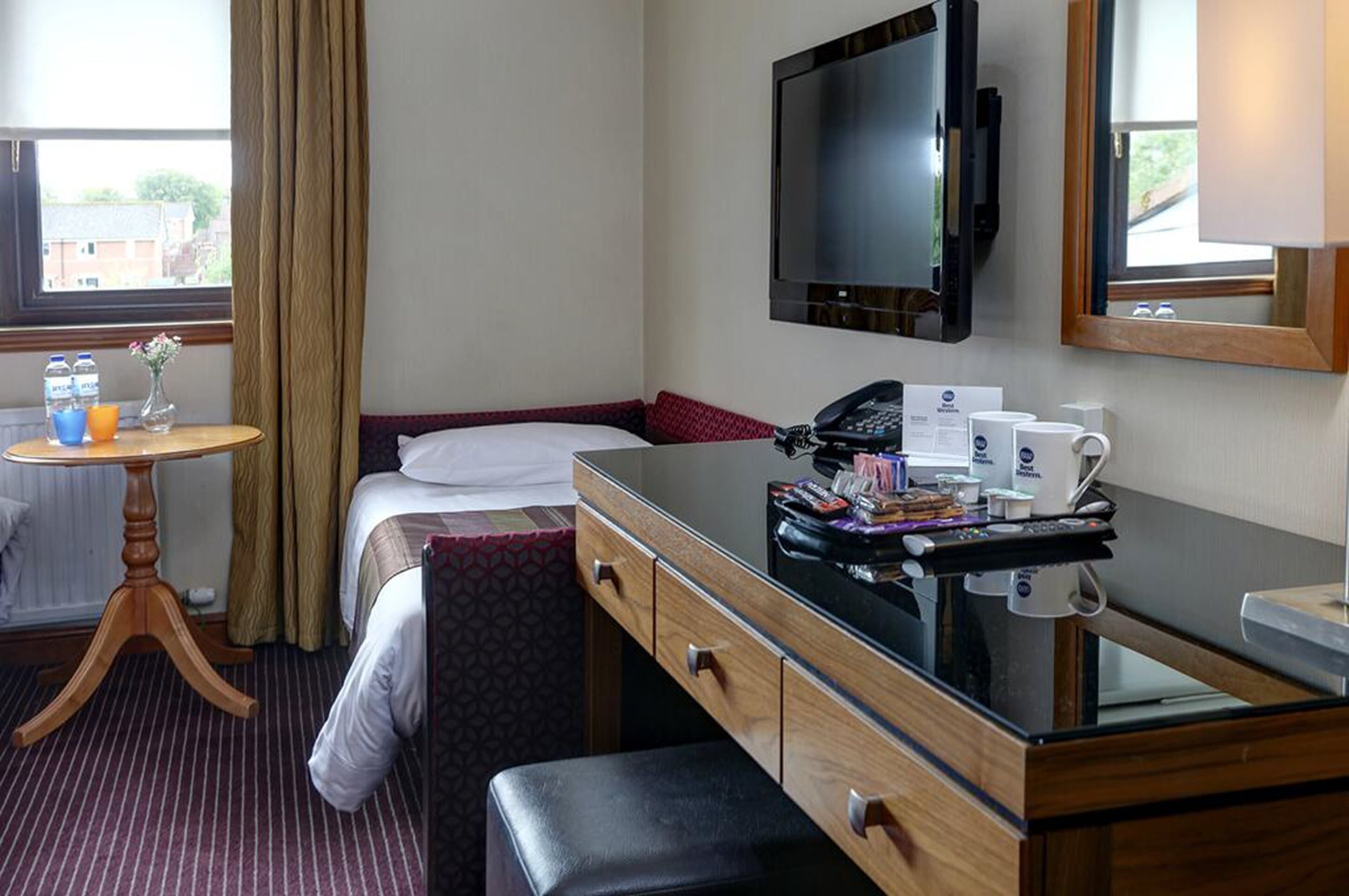 A hotel room with a bed, a desk with a telephone, a flat-screen TV, and a small table with a vase and water bottles.