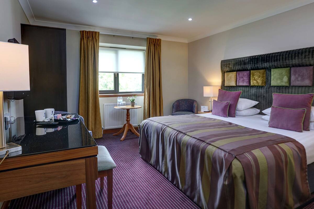 Une chambre d’hôtel comprenant un lit, un bureau avec un ordinateur, une chaise, une fenêtre avec des rideaux et un mur orné d’un panneau décoratif.