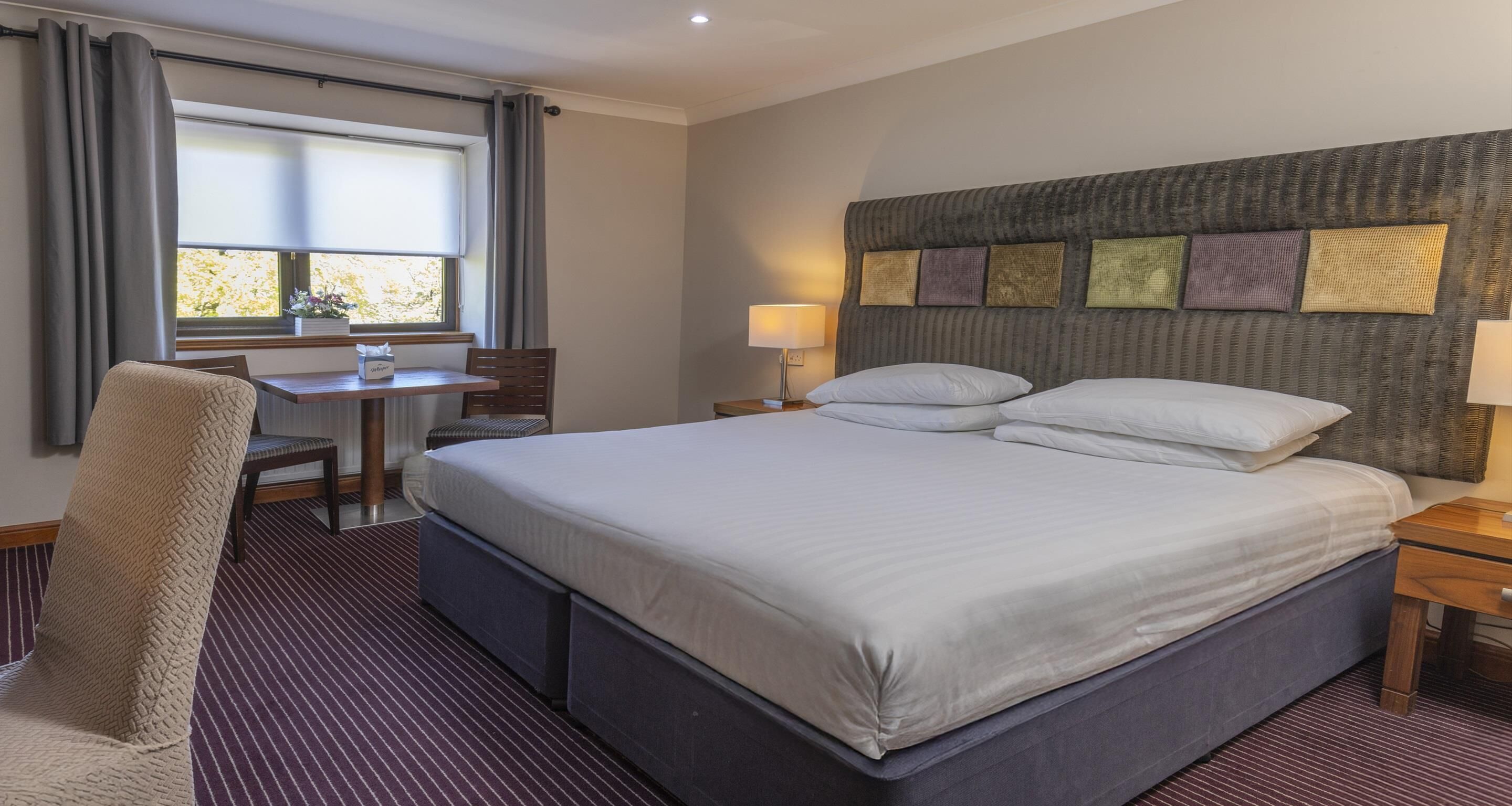 A hotel room with a large bed, a desk, and a chair. There is a window with curtains, a headboard with decorative pillows, and two bedside lamps.