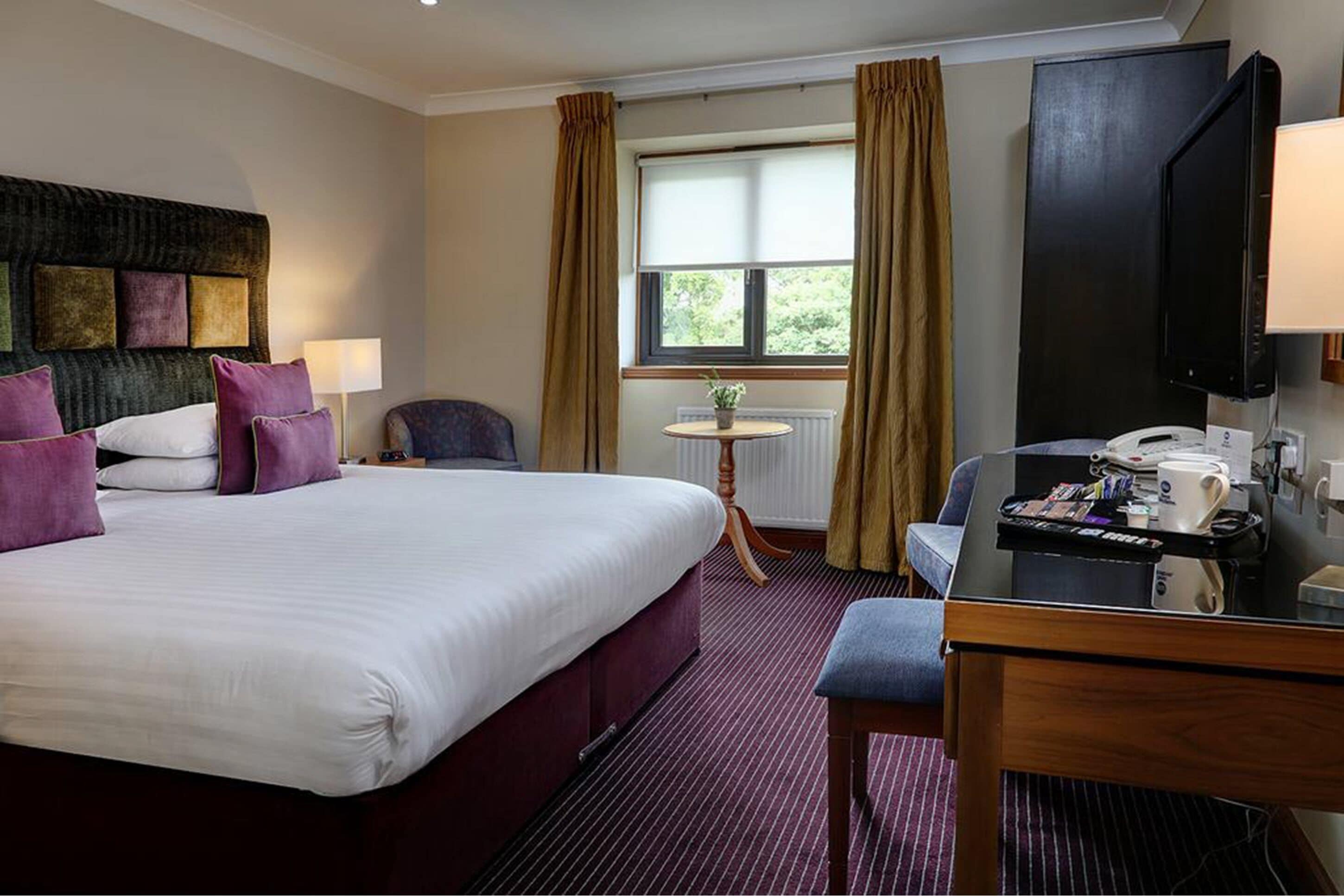 A hotel room with a large bed, a desk with a coffee maker, a television, and a window with curtains.