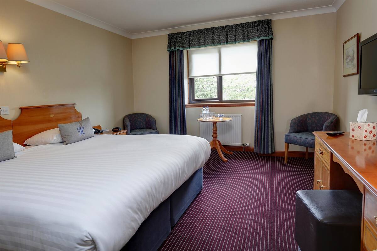 Une chambre d’hôtel avec un grand lit, deux chaises, un bureau, une petite table et une fenêtre avec des rideaux.