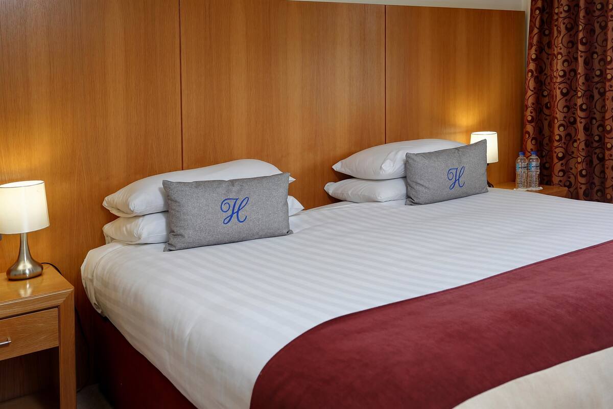 Un lit bien fait, avec une literie blanche et grise, deux oreillers ornés d’un monogramme, une tête de lit en bois, une table de chevet avec une lampe et un rideau à motifs.
