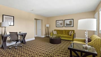 A hotel room with a patterned carpet, a green sofa, a yellow armchair, a glass coffee table, and a desk with a chair.