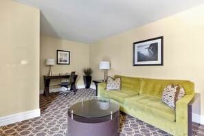 A living room with a green sofa, a glass coffee table, a desk with a chair, and two lamps. - Marin Suites Hotel (Corte Madera)