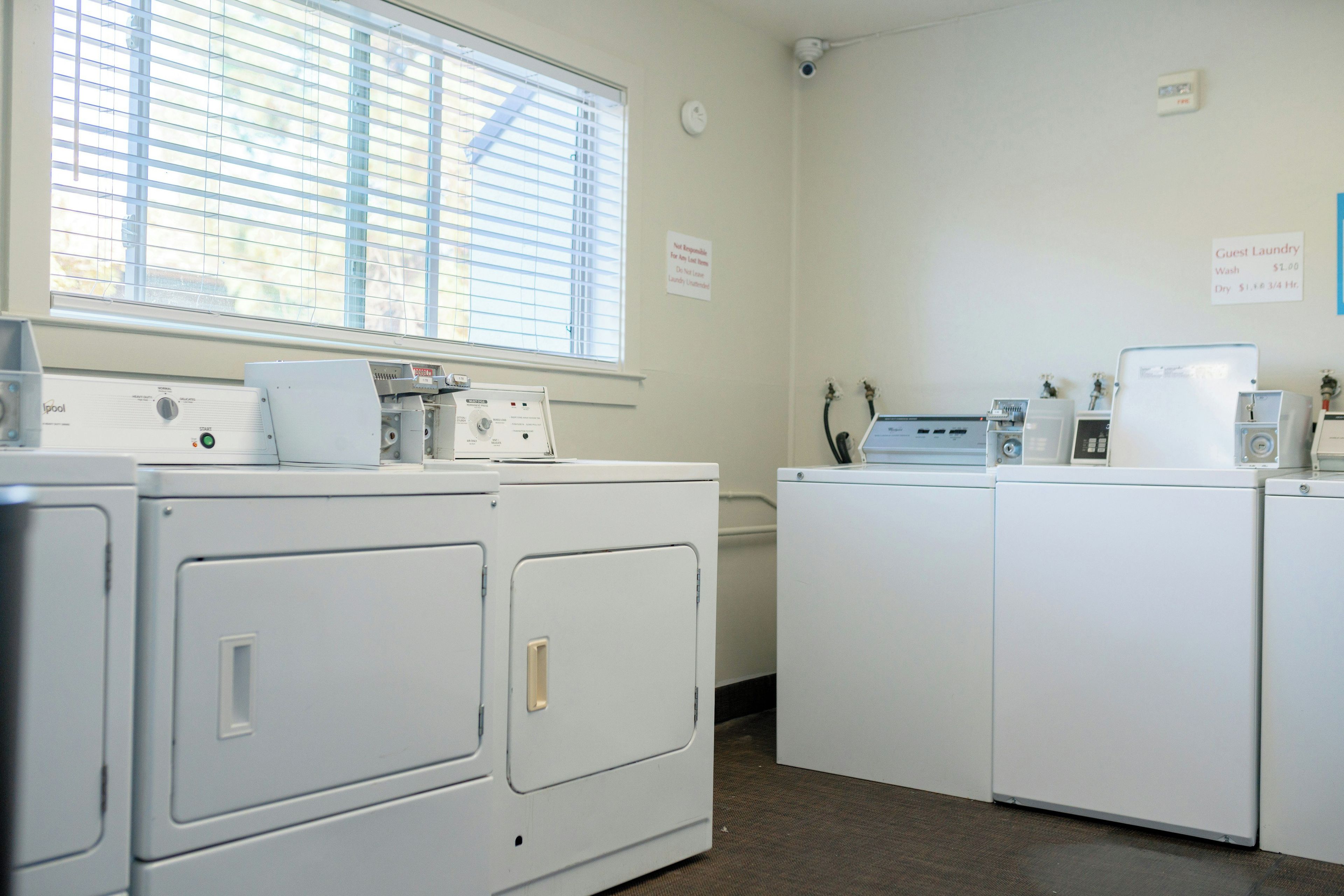 A laundry room with four white washing machines, a dryer, and a wall-mounted washing machine. There is a window with blinds, a sign indicating guest laundry services, and a thermostat on the wall.