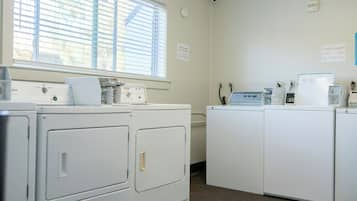 A laundry room with four white washing machines, a dryer, and a wall-mounted washing machine. There is a window with blinds, a sign indicating guest laundry services, and a thermostat on the wall.