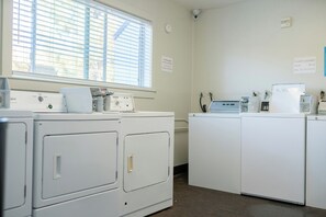 A laundry room with four white washing machines, a dryer, and a wall-mounted washing machine. There is a window with blinds, a sign indicating guest laundry services, and a thermostat on the wall. - Marin Suites Hotel (Corte Madera)