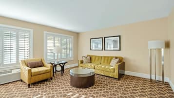 A living room with a patterned carpet, a yellow sofa, a yellow armchair, a glass coffee table, a floor lamp, and two framed pictures on the wall.