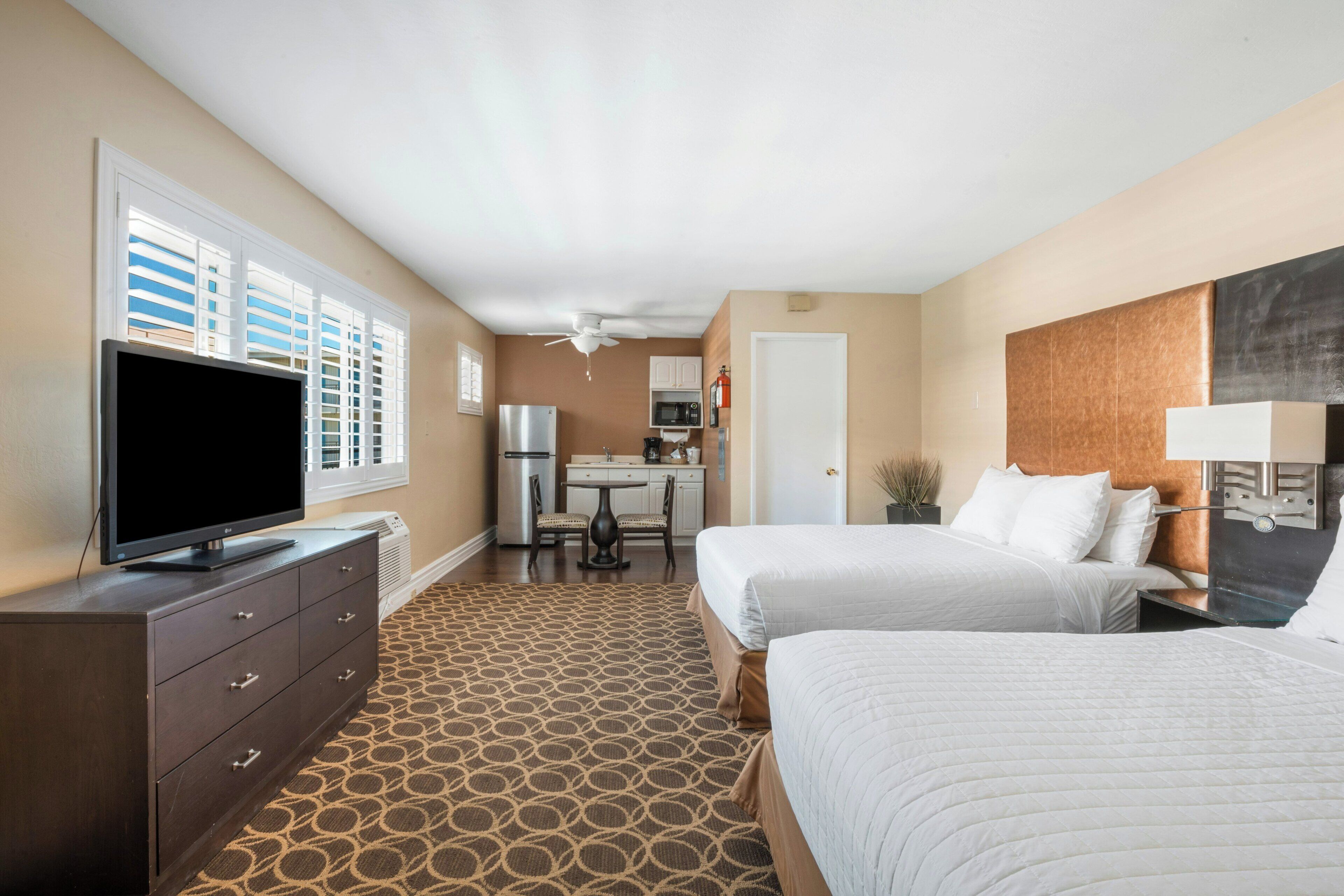 A hotel room with two beds, a TV, a kitchenette, and a ceiling fan.