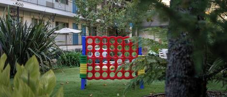 A playground with a red and yellow climbing structure, surrounded by greenery and a building with a balcony in the background.