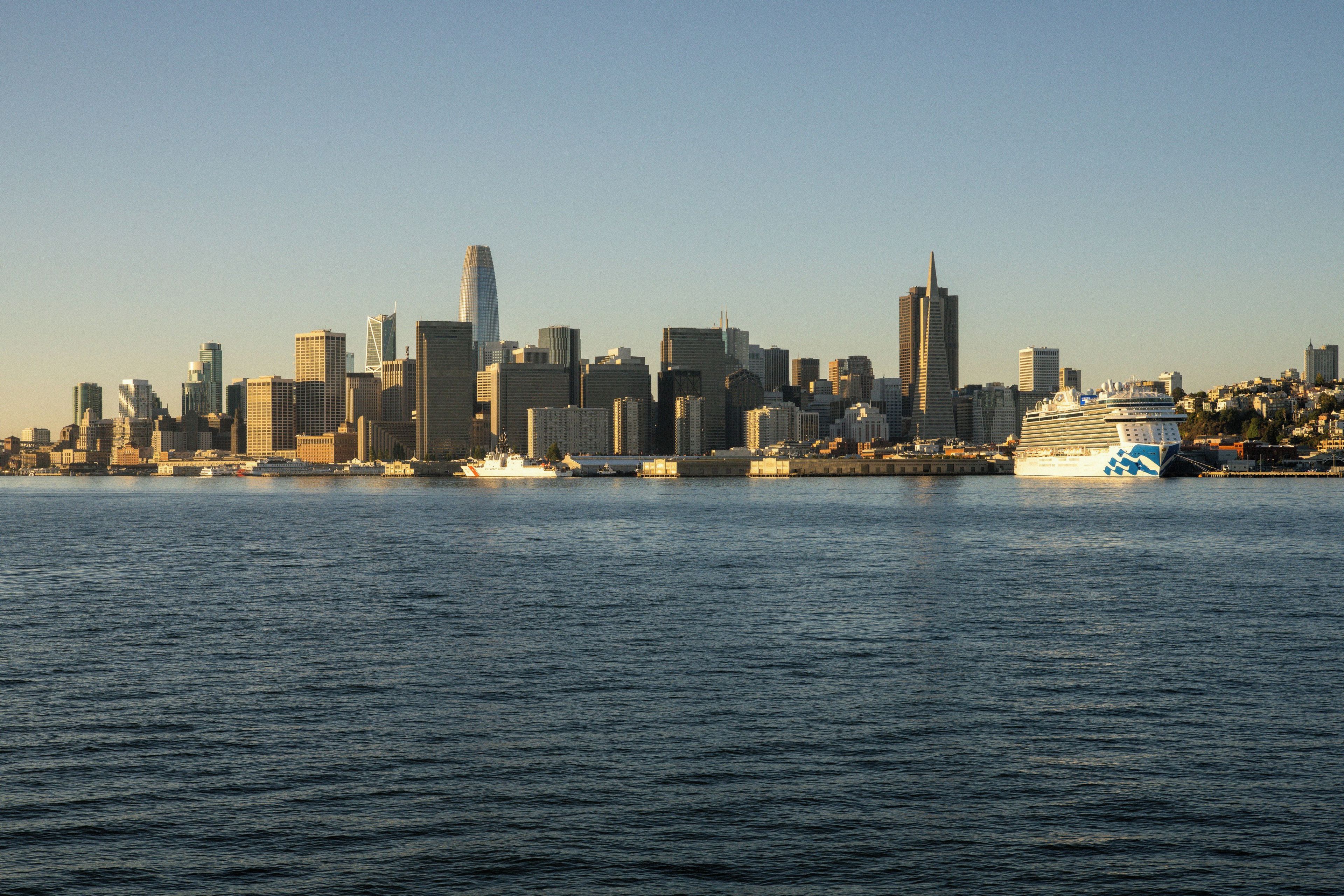 Une vue sur les toits d’une ville, avec des bâtiments modernes et un grand navire de croisière amarré au bord de l’eau.