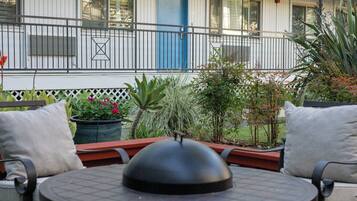 Una mesa redonda al aire libre con una cubierta tipo cúpula, rodeada por cuatro sillas con cojines. Al fondo, se observa un edificio con una puerta azul y un balcón con barandillas de metal.