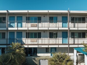 A multi-story building with balconies, a sign indicating the number 100, and a small garden area with plants. - Marin Suites Hotel (Corte Madera)