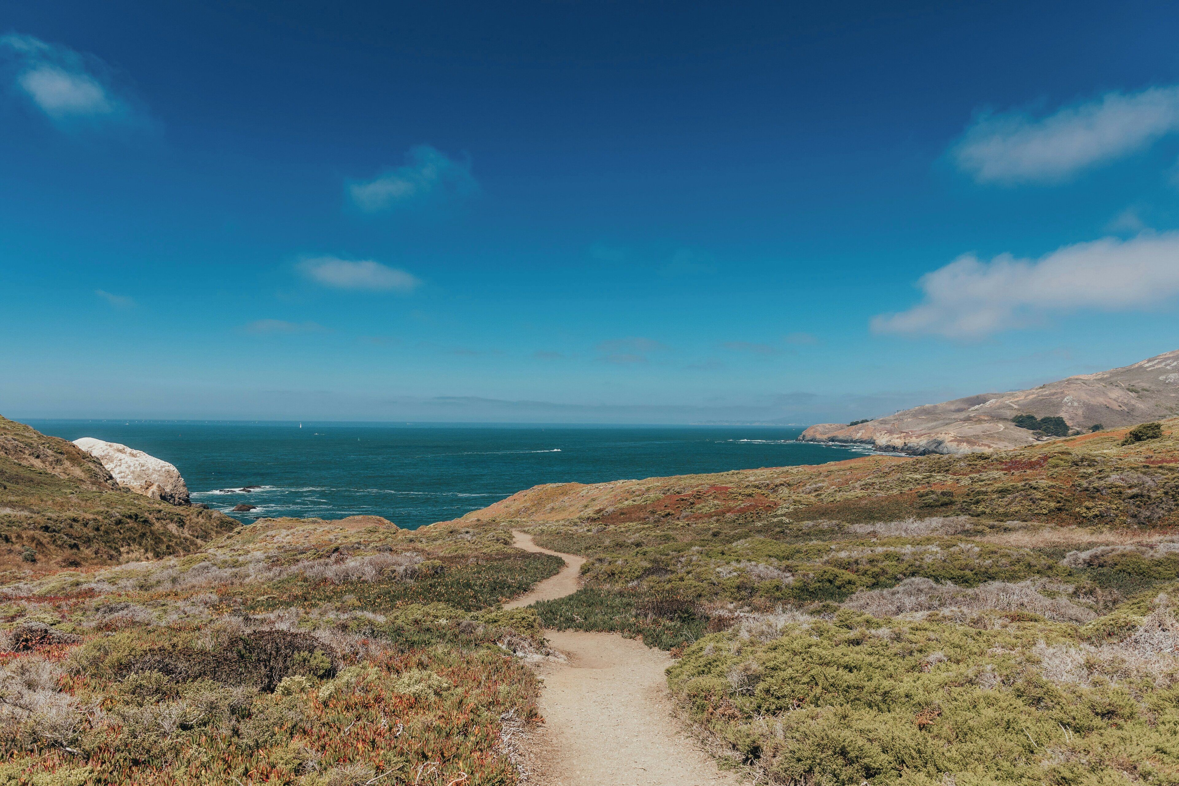 A coastal landscape with a dirt path leading to the sea, surrounded by hills and sparse vegetation.