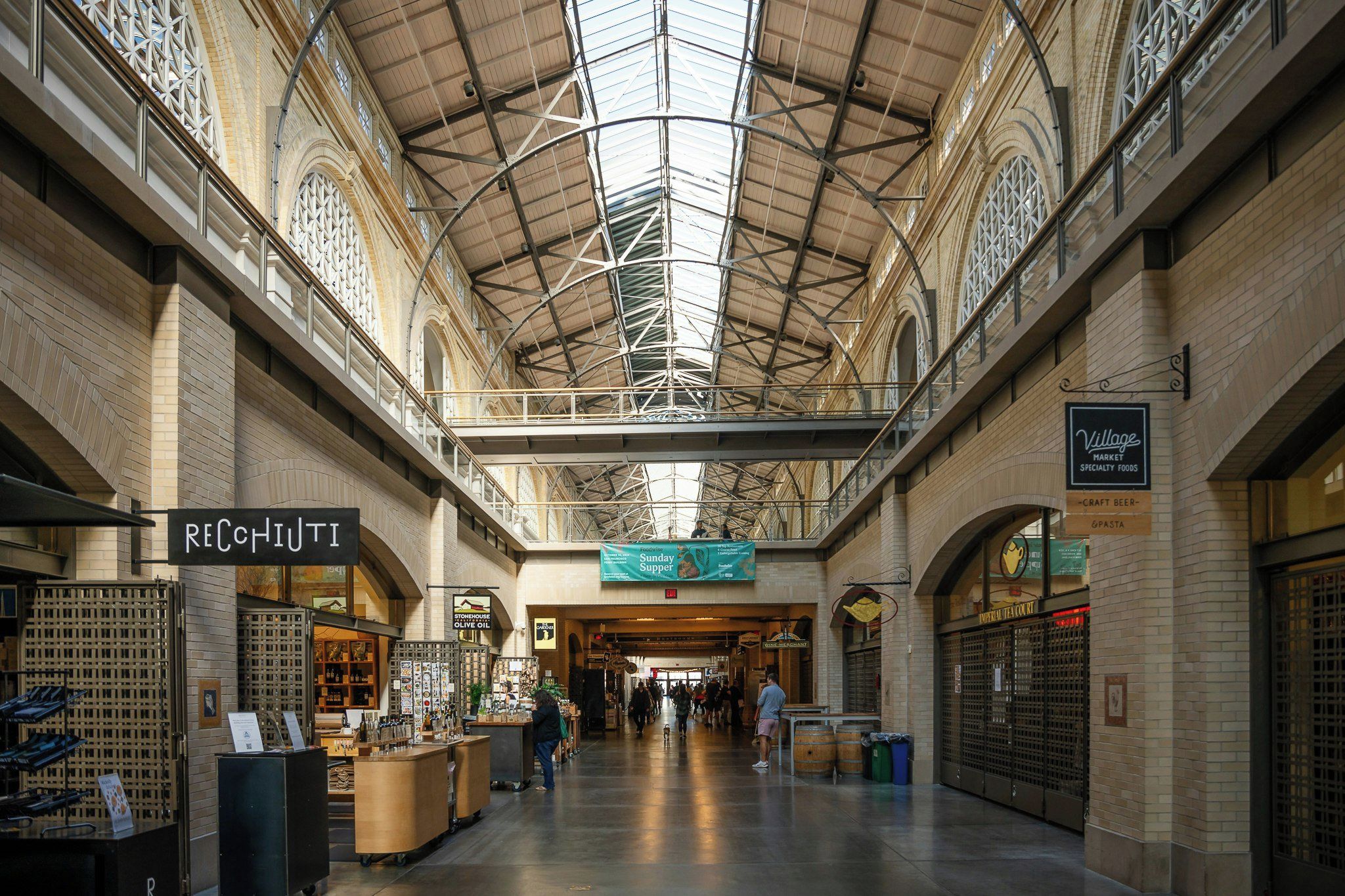 An indoor market with a high ceiling and arched windows, featuring shops and a sign for a Sunday supper.
