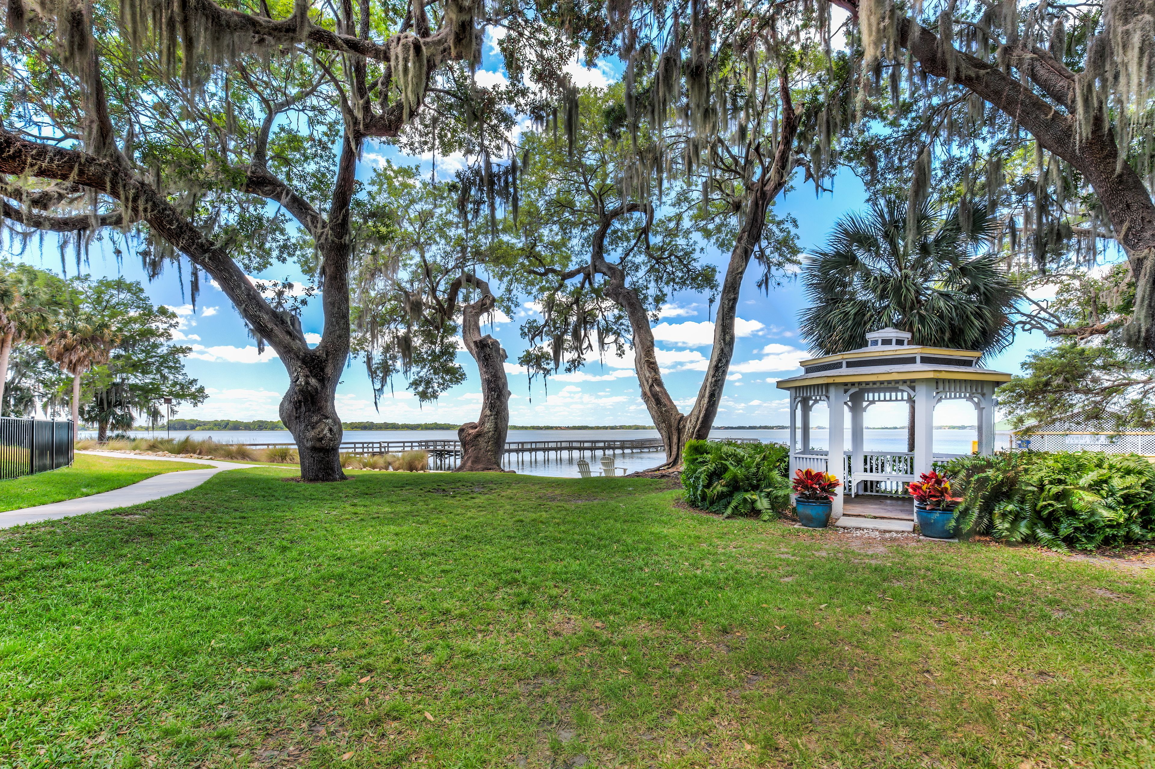 A gazebo with a white roof and blue trim, surrounded by lush greenery and a view of a lake.