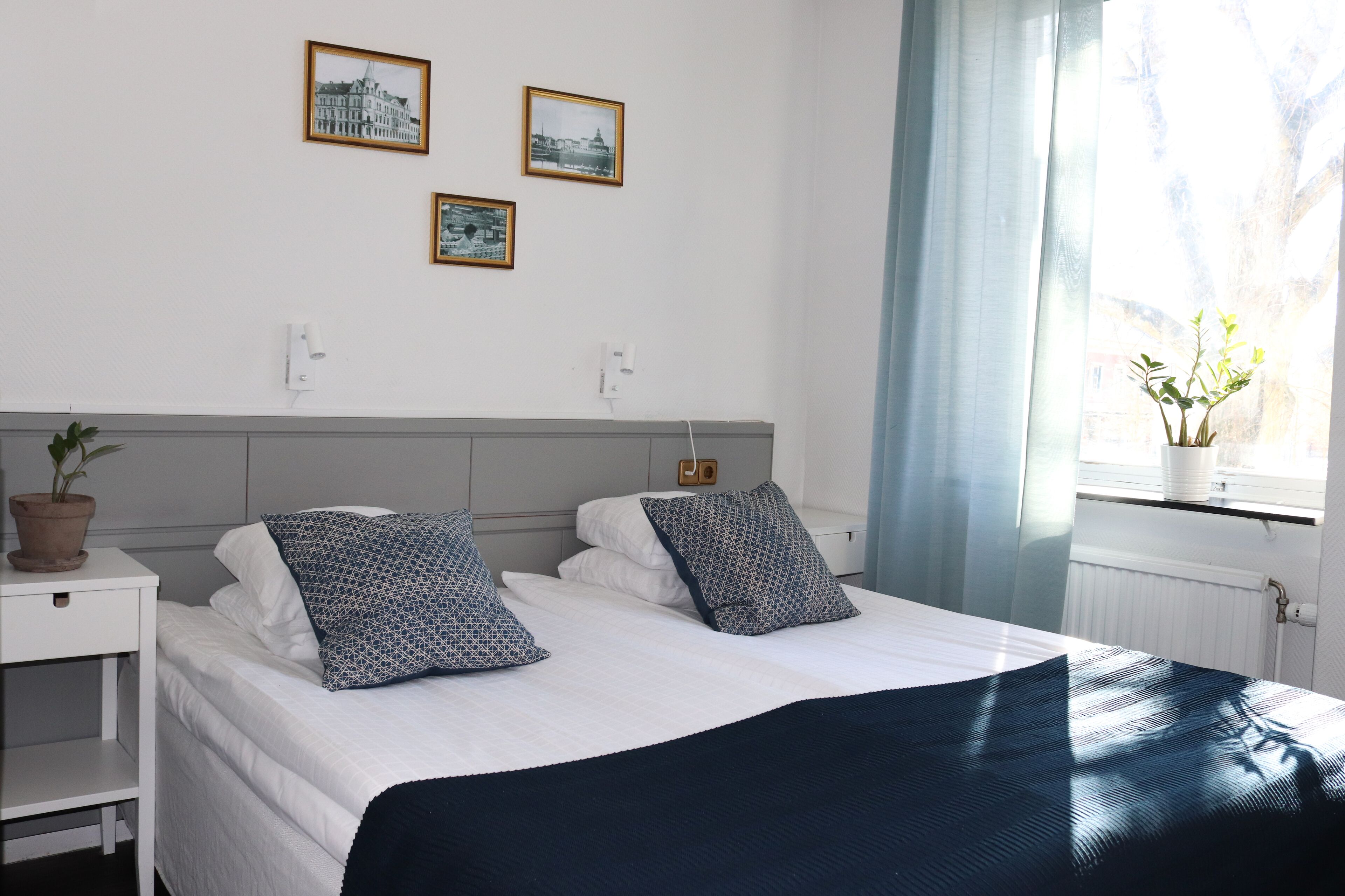 Cama bem arrumada com almofadas estampadas, uma planta na mesinha de cabeceira e janela com vista para árvores.