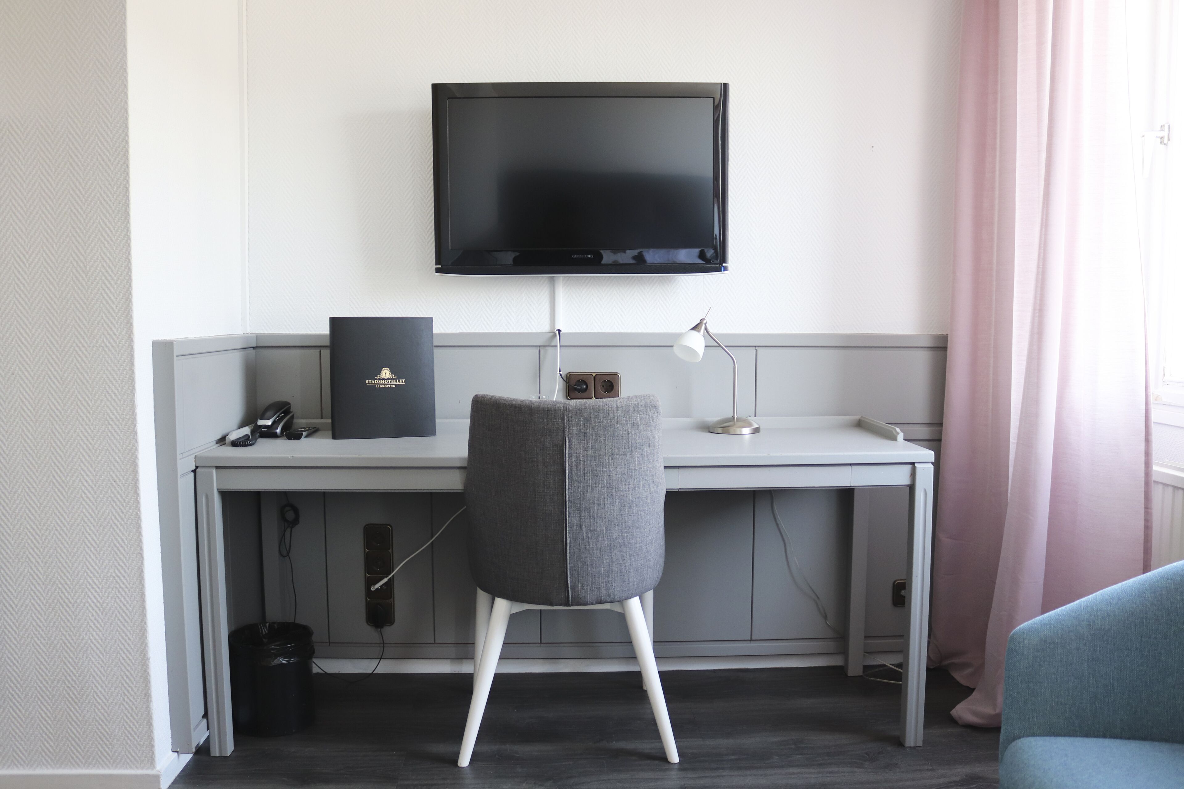 Ein modernes Hotelzimmer mit einem grau gepolsterten Sessel, einem an der Wand befestigten Flachbildfernseher, einem Schreibtisch mit Lampe und einem Fenster mit rosa Vorhängen.