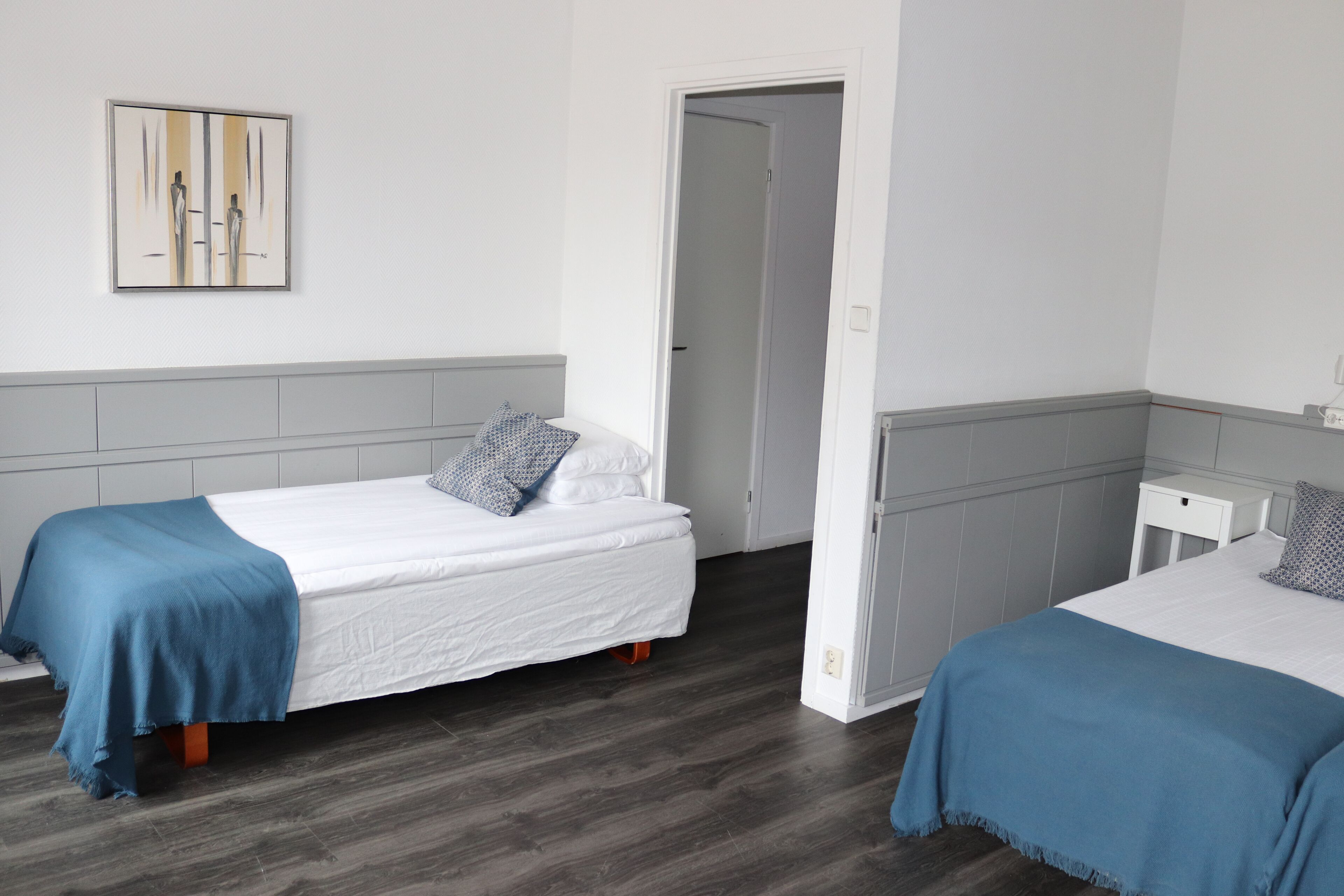 A hotel room with two single beds, a grey headboard, a white wall, and a door leading to another room.