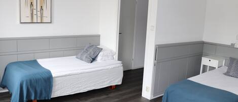 Une chambre d’hôtel avec deux lits simples, une tête de lit grise, un mur blanc et une porte donnant sur une autre pièce.