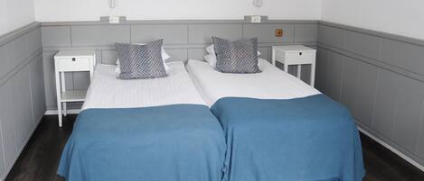 Una habitación doble con dos camas, cada una con una manta azul y almohadones estampados. Hay mesitas de noche con lámparas y un cuadro enmarcado en la pared.