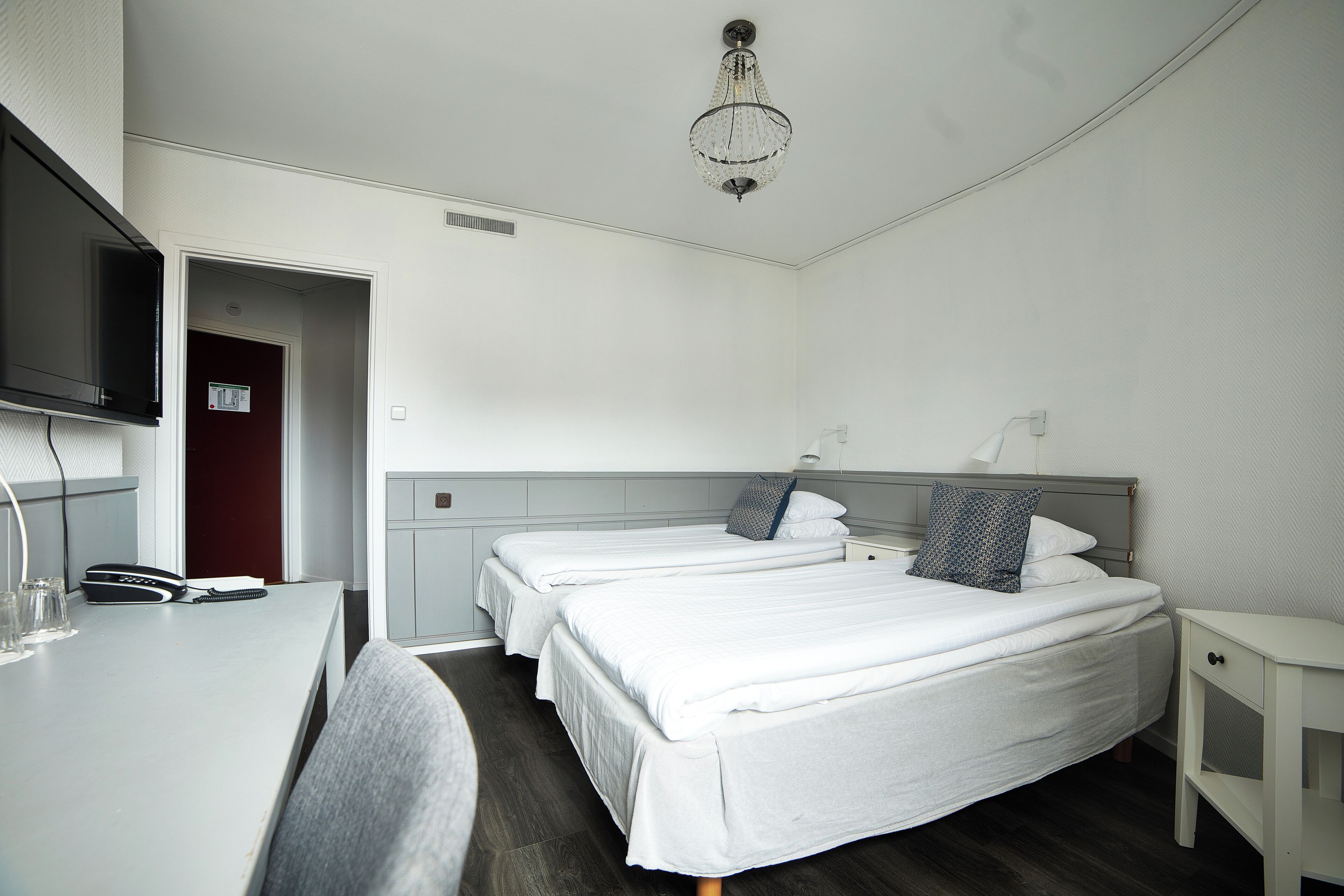 Ett hotellrum med två separata sängar, ett skrivbord med en telefon och ett par glasögon, en tv som är monterad på väggen och en kristallkrona.