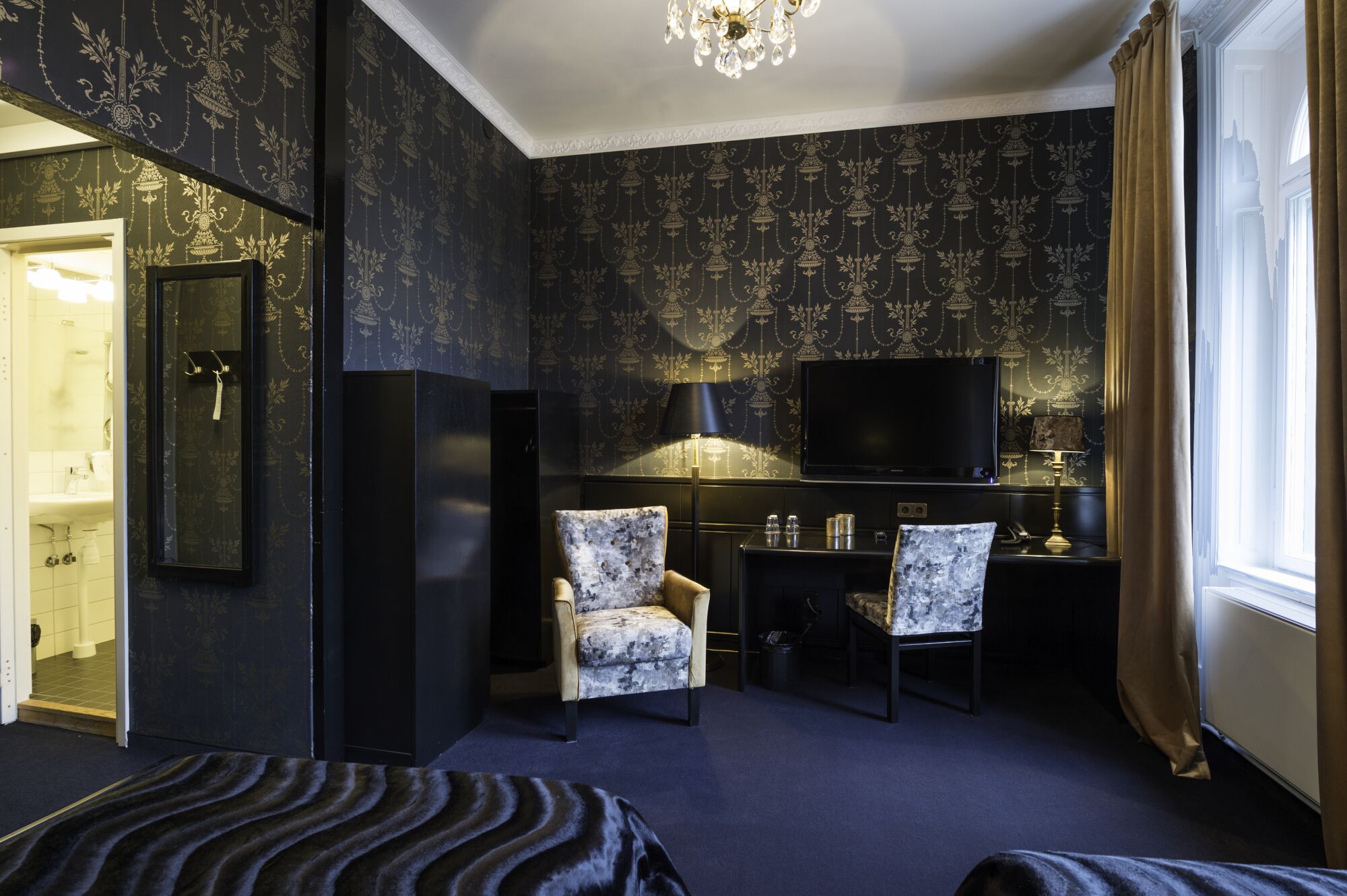 Une chambre d’hôtel avec un papier peint à motifs sombres, une télévision, deux fauteuils, un bureau avec une lampe et une grande fenêtre avec des rideaux.