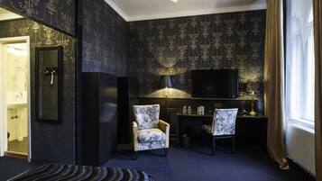 Une chambre d’hôtel avec un papier peint à motifs sombres, une télévision, deux fauteuils, un bureau avec une lampe et une grande fenêtre avec des rideaux.