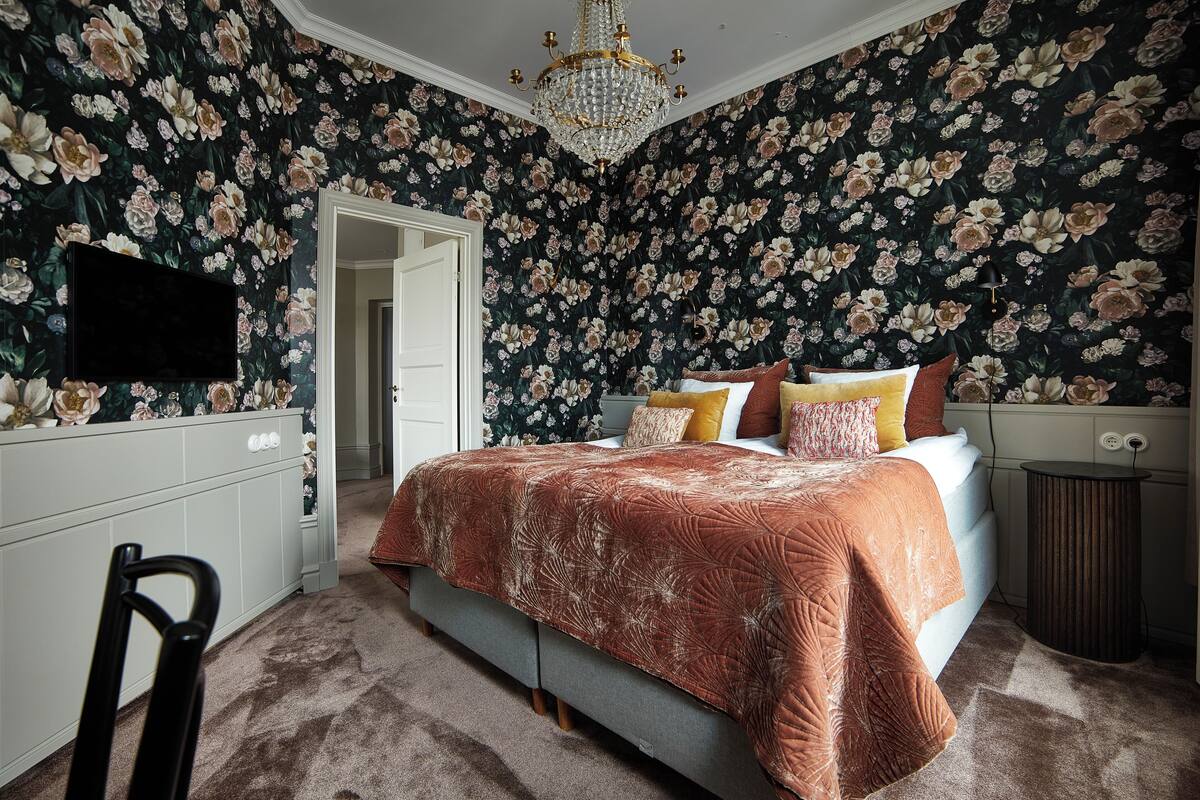 A bedroom with floral wallpaper, a bed with a patterned bedspread, a chandelier, and a television on the wall.