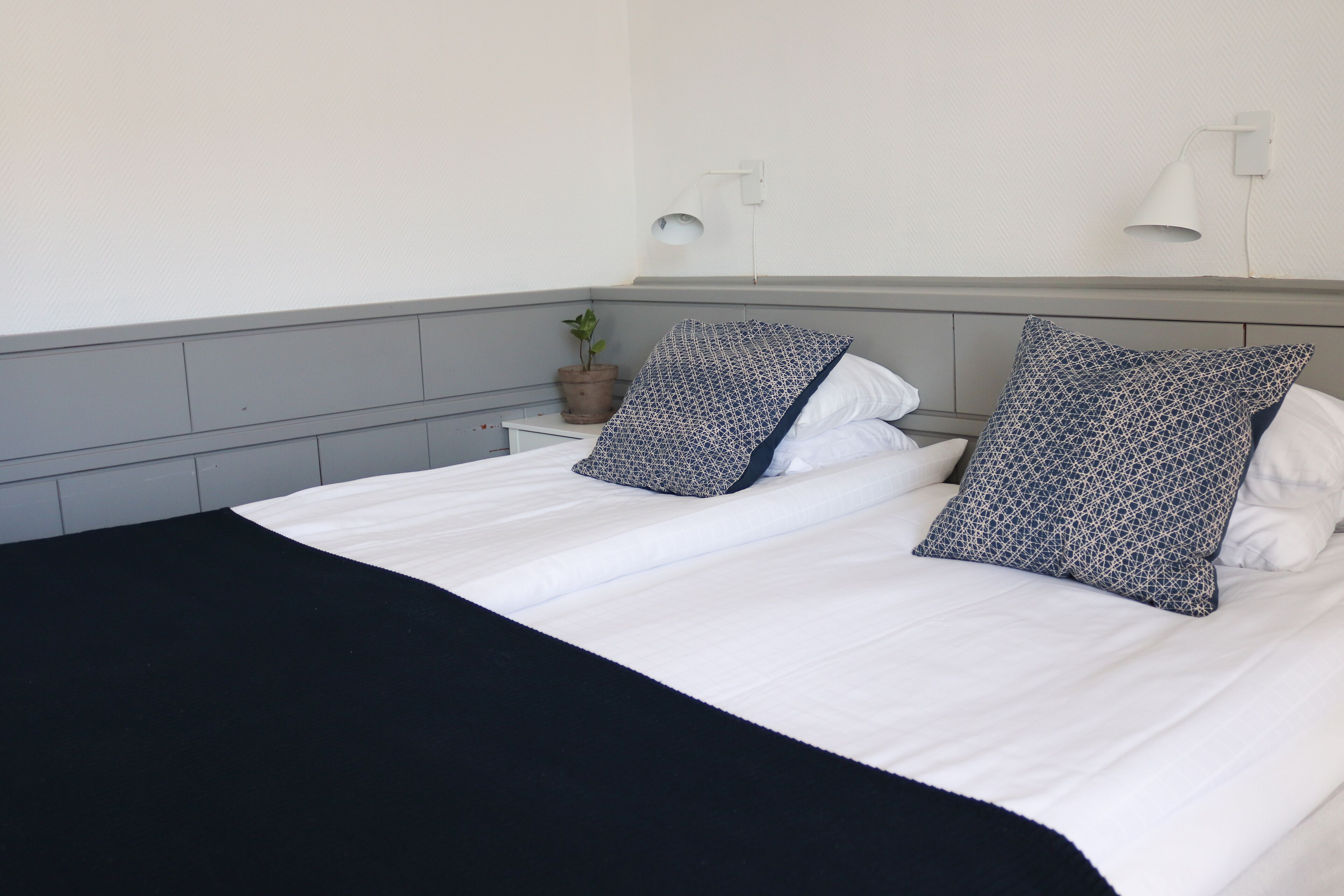 A neatly made bed with white bedding and patterned pillows, a bedside plant, and a wall-mounted lamp.
