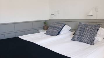 A neatly made bed with white bedding and patterned pillows, a bedside plant, and a wall-mounted lamp.