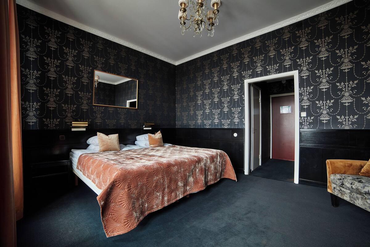 A bedroom with a large bed, dark patterned wallpaper, a mirror, and a chandelier.