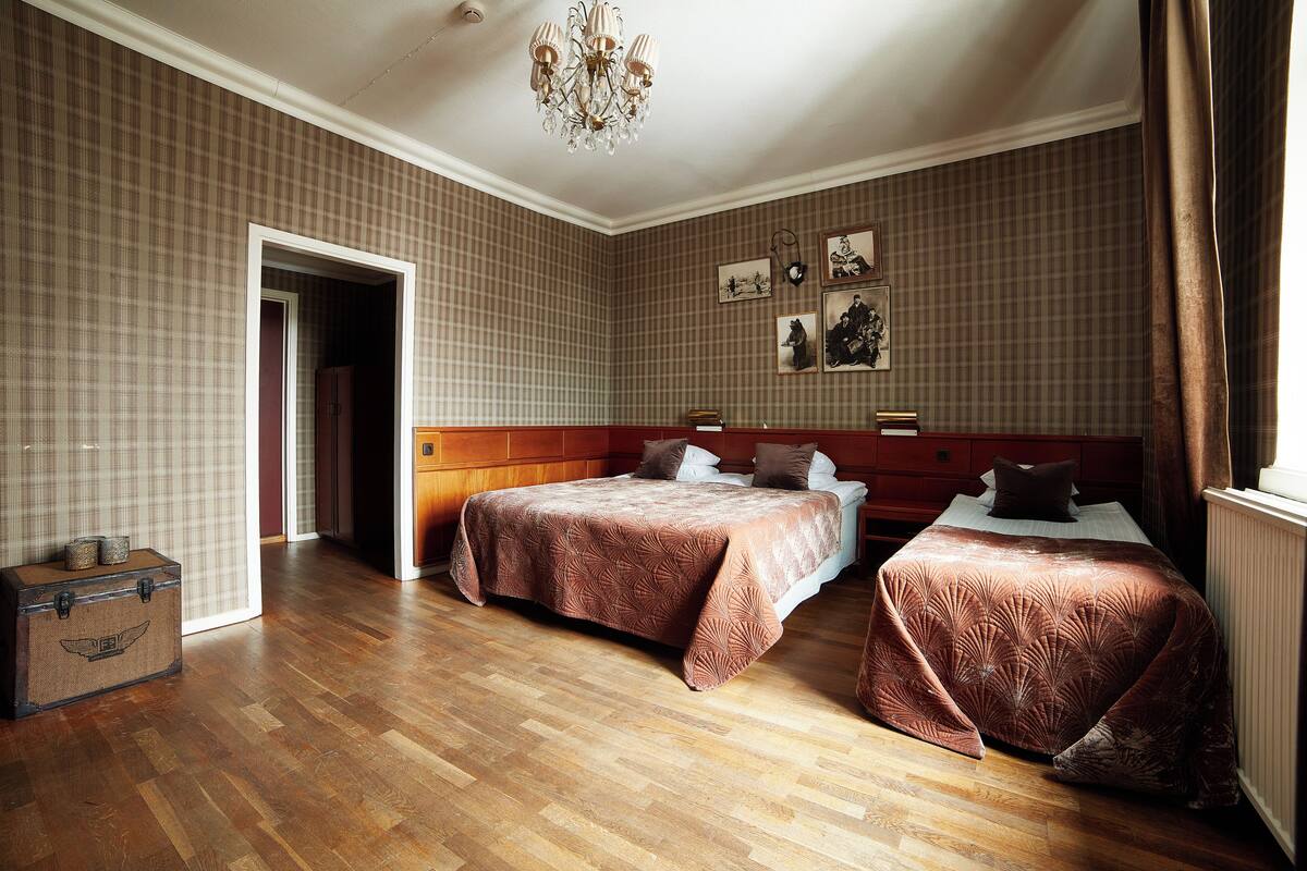 A hotel room with two beds, wooden flooring, a chandelier, and plaid wallpaper.