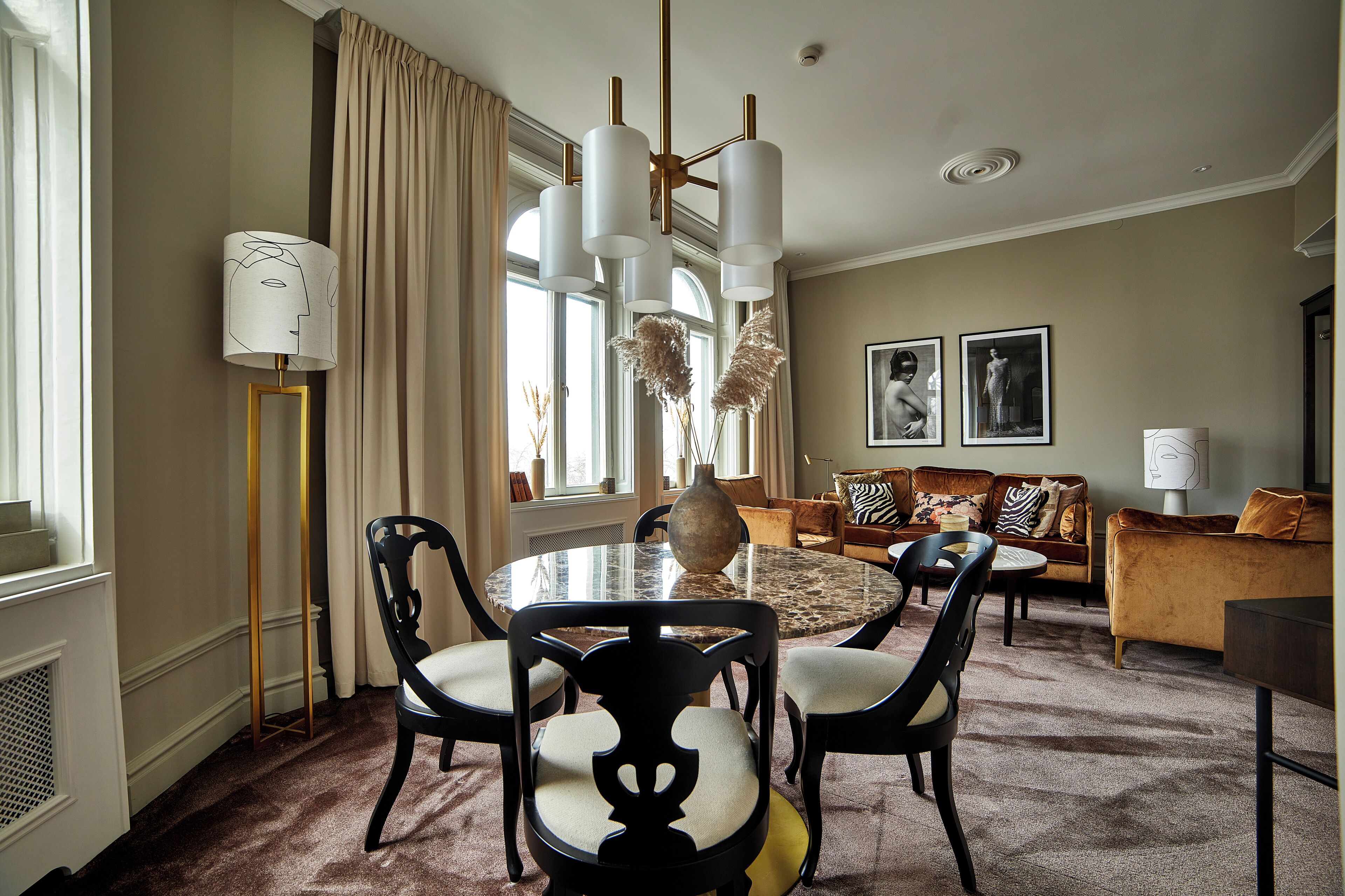 A dining area with a round table, chairs, and a vase. There is a sofa and a lamp in the background.